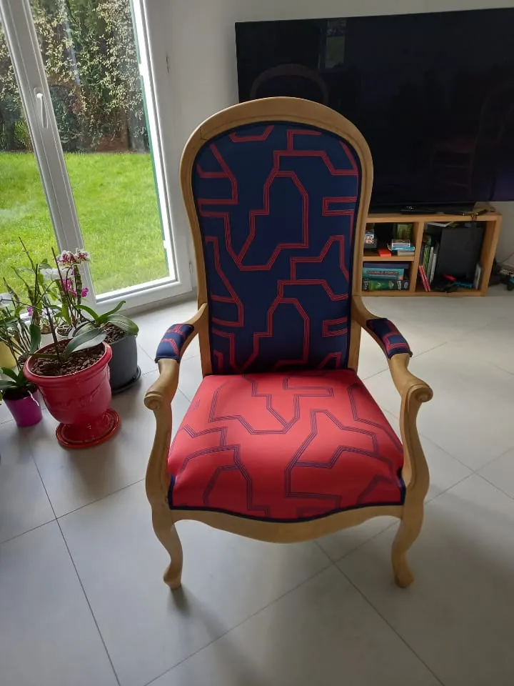 Fauteuil Voltaire en bois clair retapissé d'un tissu géométrique bleu et rouge, dans un salon lumineux